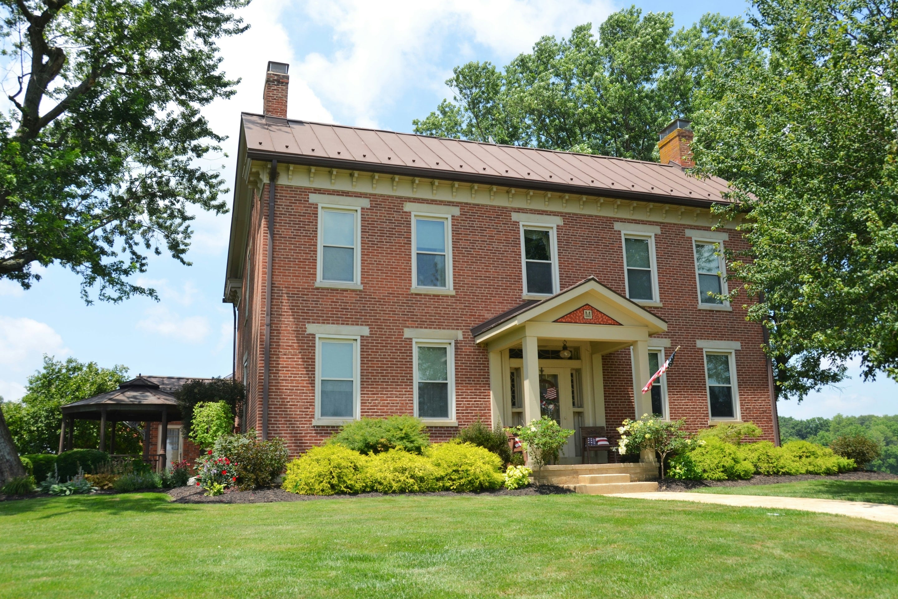 A two-story red brick house with a metal roof and a columned entry porch, behind a mown summer lawn and shade trees